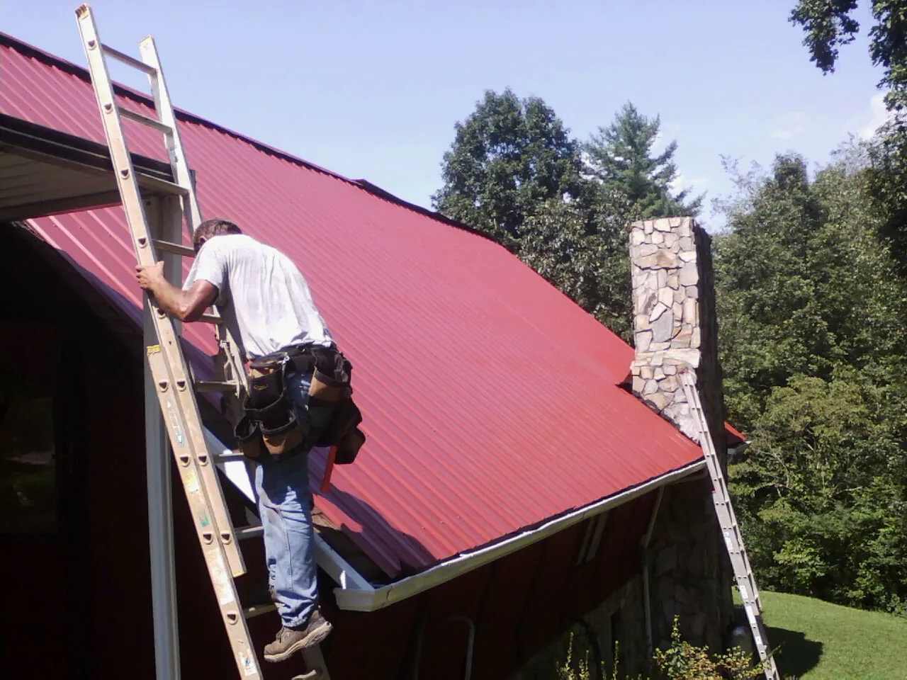 Roofer on a ladder beside a red metal roof and stone chimney — Soffit & Fascia Repair by Roofing Brothers in Sumava Resorts
